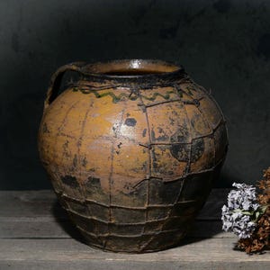 Pode incluir: Um pote de cerâmica grande e redondo, de cor amarelo-mostarda, com acabamento escuro e desgastado e uma rede de arame. Ao lado, há um pequeno jarro de cerâmica preto fosco e um ramo de flores secas castanhas e cinzentas.