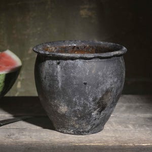 May include: A dark grey, weathered ceramic pot with a wide, scalloped rim sits next to a halved watermelon with a red interior and green rind. A small knife with a wooden handle lies horizontally on the surface in front of the pot and fruit.