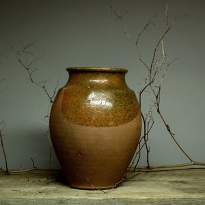 May include: A brown ceramic vase with a speckled glaze sits on a weathered wooden surface. Dried branches are arranged around the vase.