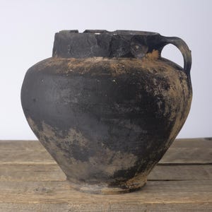 May include: A rustic, bulbous ceramic vessel with a dark, matte black finish and visible weathered, earthy brown undertones. The pot features a wide, textured rim, a single curved handle on the side, and a tapered base with a worn, aged appearance.