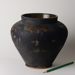 Könnte beinhalten: Eine große, dunkel gefärbte Keramikvase mit strukturierter Oberfläche und weiter Öffnung. Die Vase hat einen runden Körper und einen leicht ausgestellten Boden. Ein grüner Bleistift liegt auf der Oberfläche neben der Vase.