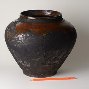 May include: A large, dark ceramic pot with a wide body and a narrow neck. The pot is primarily black with areas of brown and silver-gray. The texture appears rough and aged. An orange pencil rests in front of the pot.