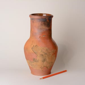 Puede incluir: Un jarrón alto de color terracota con una base ancha y un cuello estrecho. El jarrón tiene un aspecto envejecido con zonas de pintura descascarillada y decoloración. Un lápiz naranja descansa sobre la superficie cerca del jarrón.