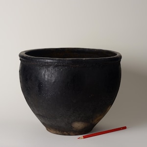 May include: A large, black ceramic pot with a wide opening and rounded body. The pot has a textured surface and a dark, matte finish. A red pencil rests on the surface near the pot, providing a size reference. The pot is likely for decorative or functional use.