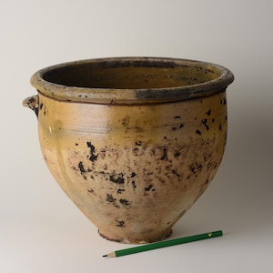 Può includere: Un grande vaso in ceramica in stile antico con un'ampia apertura e due piccole maniglie. Il vaso ha una finitura screziata gialla e marrone con macchie scure. Una matita verde è appoggiata sulla superficie vicino al vaso.