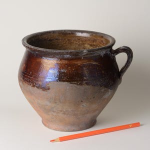 Puede incluir: Una olla de cerámica marrón con asa. La olla tiene una abertura ancha y un cuerpo redondeado. Un lápiz naranja descansa sobre la superficie junto a la olla.