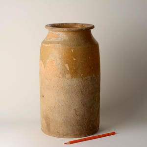 May include: A cylindrical stoneware jar with a wide mouth and a thick, rolled rim. The upper portion features a smooth, glossy tan glaze, while the lower section has a rough, matte, unglazed beige texture. An orange pencil lies in the foreground.