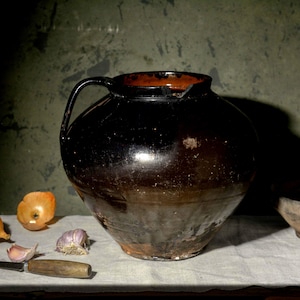 Può includere: Una grande brocca in ceramica smaltata marrone scuro con un solo manico, posizionata accanto a una cipolla gialla, un bulbo d'aglio, un piccolo coltello con manico in legno e una ciotola in ceramica poco profonda e usurata con il bordo incrinato.