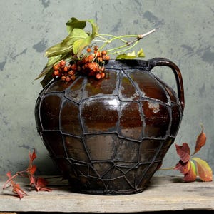 May include: A dark brown ceramic vase with a woven wire design, filled with red berries and green leaves. The vase has a wide mouth and two handles.