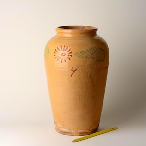 Può includere: Un alto vaso in ceramica gialla di forma cilindrica con un'apertura ampia e un bordo spesso. L'esterno presenta motivi dipinti a mano a forma di sole marrone e disegni di foglie verdi. Una matita gialla è posta alla base come riferimento.