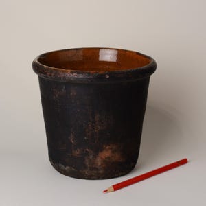 May include: A dark, weathered ceramic pot with a wide rim and a reddish-brown interior. The exterior is a mottled black with hints of brown. A red pencil rests on the surface next to the pot. Suitable for plants.