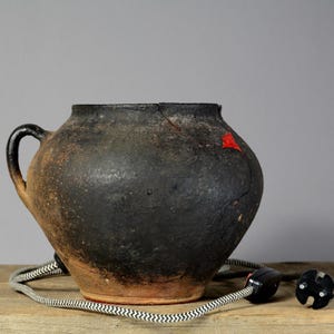 May include: A rustic, black ceramic pot with a single handle, featuring a red patch and a crack. A black and white striped cord with a black plug is draped in front of the pot. The pot is on a wooden surface.