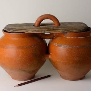 May include: A double-chambered, terracotta-colored ceramic pot with a wooden lid and handle. The pot has a rustic appearance with visible imperfections. A brown pencil rests in front of the pot.