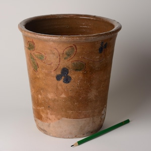 May include: A tall, cylindrical terracotta pot with a wide rim. The pot is decorated with green and blue floral designs and a reddish-brown vine. A green pencil rests on the surface next to the pot.