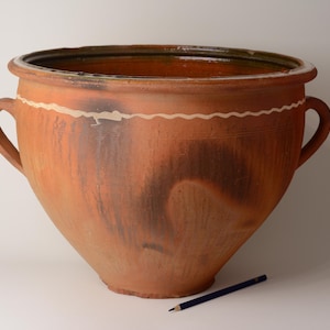 May include: Large, antique terracotta pot with two handles. The pot has a wide mouth and a rounded body, with a wavy white painted design around the top. Darker brown and black markings add to the aged appearance. A blue pencil rests on the surface.