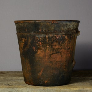 Può includere: Un grande vaso di terracotta marrone scuro con una finitura rustica usurata. Il vaso ha un'ampia apertura rotonda e un labbro leggermente svasato. La superficie è irregolare e mostra segni di età e utilizzo.