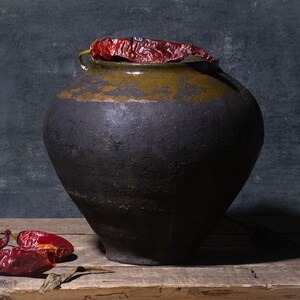 May include: A dark, ceramic jar with a wide base and a narrow opening, adorned with a dried red chilli pepper. Several dried red chilli peppers are scattered on a wooden surface in front of the jar. The background is a dark, textured wall.