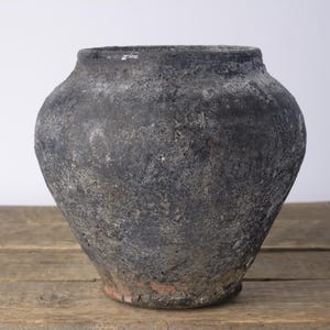 May include: A round, wide-mouthed vessel with a heavily textured, dark charcoal-gray exterior. The surface displays a weathered, matte finish with subtle variations in tone, revealing hints of reddish-brown clay near the narrow, circular base.