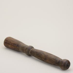 May include: A weathered, wooden pestle with a cylindrical handle and rounded ends. The pestle is a dark brown color, showing signs of wear and age. It is designed for grinding ingredients in a mortar, a tool commonly used in cooking.