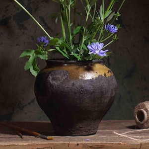 May include: A dark brown ceramic vase with a golden-brown rim, containing green foliage and purple flowers. The vase is on a wooden surface, with paintbrushes and twine in the foreground. The image has a vintage, natural aesthetic, with a focus on the vase.