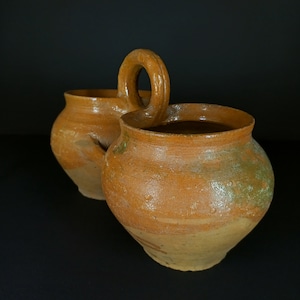May include: Two brown clay pots with a handle on one of the pots. The pots are unglazed and have a rustic appearance.