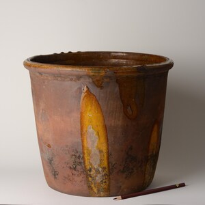 May include: A large, cylindrical brown ceramic crock featuring a thick, dark rim and vertical, elongated yellow-orange glaze drips running down the exterior surface. A thin, dark-colored pencil is positioned horizontally at the base for scale.
