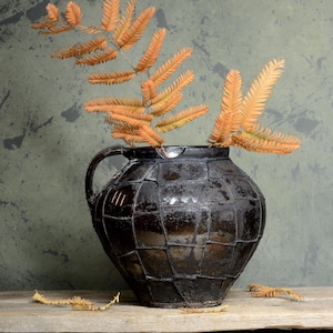 Peut inclure: Un vase en céramique noire avec une surface texturée, décoré d'un motif géométrique. Le vase est rempli de feuilles de fougère brunes séchées.