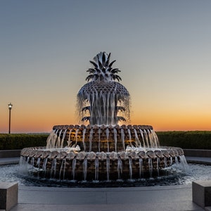 May include: A large, ornate fountain shaped like a pineapple with cascading water, surrounded by lush greenery and palm trees. The fountain is set against a backdrop of a vibrant orange and pink sunset.