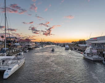May include: A scenic harbor waterway at sunset featuring multiple docked white motorboats and catamarans. The sky displays hues of orange and pink clouds above wooden piers and waterfront buildings lining both sides of the calm, dark water.