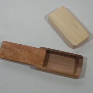 以下が含まれることがあります： 2種類の木材で作られた、スライド式の蓋が付いた長方形の木箱。蓋は明るい色で、ベースとスライド部分は濃い色の木材です。箱は開いていて空です。