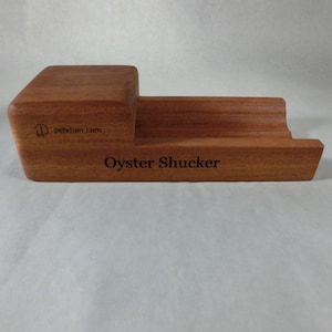 May include: A wooden oyster shucker with a natural wood grain finish. The tool has a rectangular base with a curved section and a raised platform. The text "Oyster Shucker" and "petehan.com" are visible.