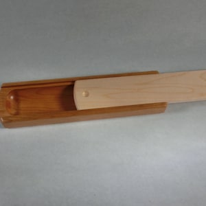 Puede incluir: Una caja de madera con tapa corredera. La caja está hecha de dos tipos de madera: una madera marrón rojiza más oscura para la base y una madera más clara y natural para la tapa. La tapa está parcialmente abierta.