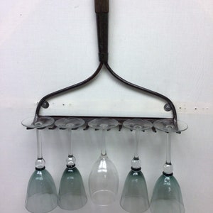 May include: A metal wine glass rack shaped like a rake with six wine glasses hanging from it. Five of the glasses are tinted green and one is clear.