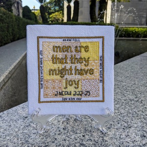 May include: A white cross-stitch embroidery with a yellow and gold design. The text reads "And men are that they might have joy. 2 Nephi 2:22-25. Adam fell that men might be."