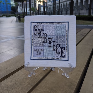 May include: A white cross-stitch embroidery with blue and black thread. The embroidery features the word "SERVICE" in a stylized font. The text "WHEN YE ARE IN THE SERVICE OF YOUR FELLOW BEINGS YE ARE ONLY IN THE SERVICE OF YOUR GOD MOSIAH 2:17" is stitched around the word "SERVICE".