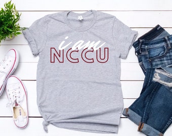 Può includere: Una maglietta grigia con il testo "i am NCCU" in lettere bianche con un contorno marrone. La maglietta è stesa su una superficie di legno bianco con un paio di scarpe da ginnastica bianche e un paio di jeans blu.