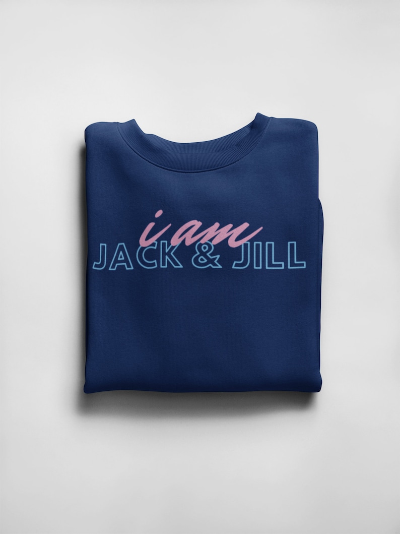 May include: A navy blue sweatshirt with the text "i am JACK & JILL" in pink and light blue. The sweatshirt is folded neatly on a white surface. The text is in a script font.