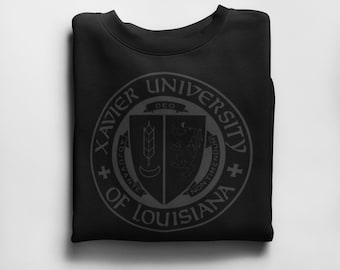 May include: Black sweatshirt with a white graphic of the Xavier University of Louisiana seal. The seal features a shield with a wheat stalk, a crescent moon, and a lion.