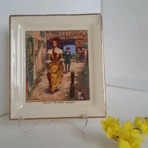 May include: A white ceramic square plate with a gold trim features a vintage illustration of a woman in a yellow dress walking down a cobblestone alley. The text "Sally in our Alley" is printed below the image.