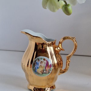 May include: A gold-plated porcelain creamer with a white floral design. The creamer has a round body with a handle and a spout. The floral design features a group of women in a garden setting.