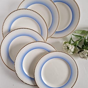 May include: Set of six white ceramic dessert plates with a blue and gold trim. The plates are stacked on top of each other.