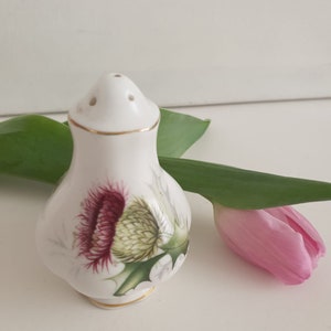 May include: White ceramic salt shaker with a gold trim and a floral design featuring a purple and green thistle. The shaker has three holes on the top for dispensing salt.