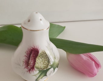 Peut inclure: Saliere en céramique blanche avec une bordure dorée et un motif floral représentant un chardon violet et vert. Le salière a trois trous sur le dessus pour dispenser le sel.