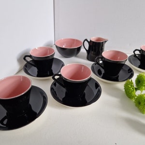 May include: A vintage tea set featuring black teacups with pink interiors, matching saucers, a small bowl, and a creamer jug. The set is arranged on a white surface. The cups and bowl have a pink interior and black exterior.