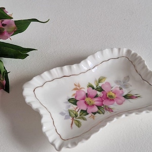 Può includere: Un piatto rettangolare in ceramica bianca con bordo smerlato e rifiniture dorate. Presenta un motivo floreale con fiori rosa e foglie verdi. Un mazzo di fiori rosa con foglie verdi è accanto.