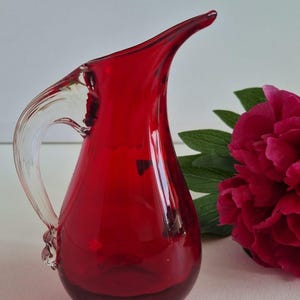 Könnte beinhalten: Eine leuchtend rote Glaskaraffe mit einem Griff aus klarem Glas. Die Karaffe hat einen gebogenen Ausguss und einen abgerundeten Boden. Eine tiefrosa Blume und grüne Blätter befinden sich im Hintergrund. Diese dekorative Karaffe ist ideal zum Servieren von Getränken.