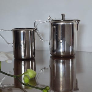 May include: A polished stainless steel creamer and a matching teapot with a hinged lid. Both vessels feature cylindrical bodies, curved handles, and integrated spouts. The metallic surfaces are highly reflective, showing clear highlights.