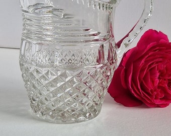 May include: A clear glass pitcher with a diamond pattern and a curved handle. The pitcher is sitting on a white surface next to a pink rose.