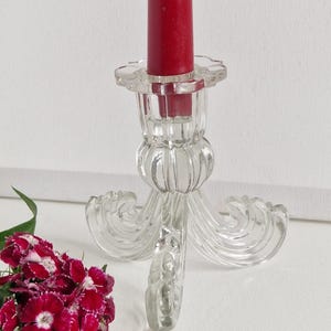 Könnte beinhalten: Ein klarer Glas-Kerzenhalter mit geriffeltem Design und drei verzierten Beinen. Der Halter ist leer und hat eine rote Kerze oben drauf.
