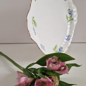 May include: A white, scalloped-edge serving dish with a gold rim, adorned with blue floral and green leaf accents. Pink and purple flowers with green leaves are in the foreground, creating a delicate, elegant composition.